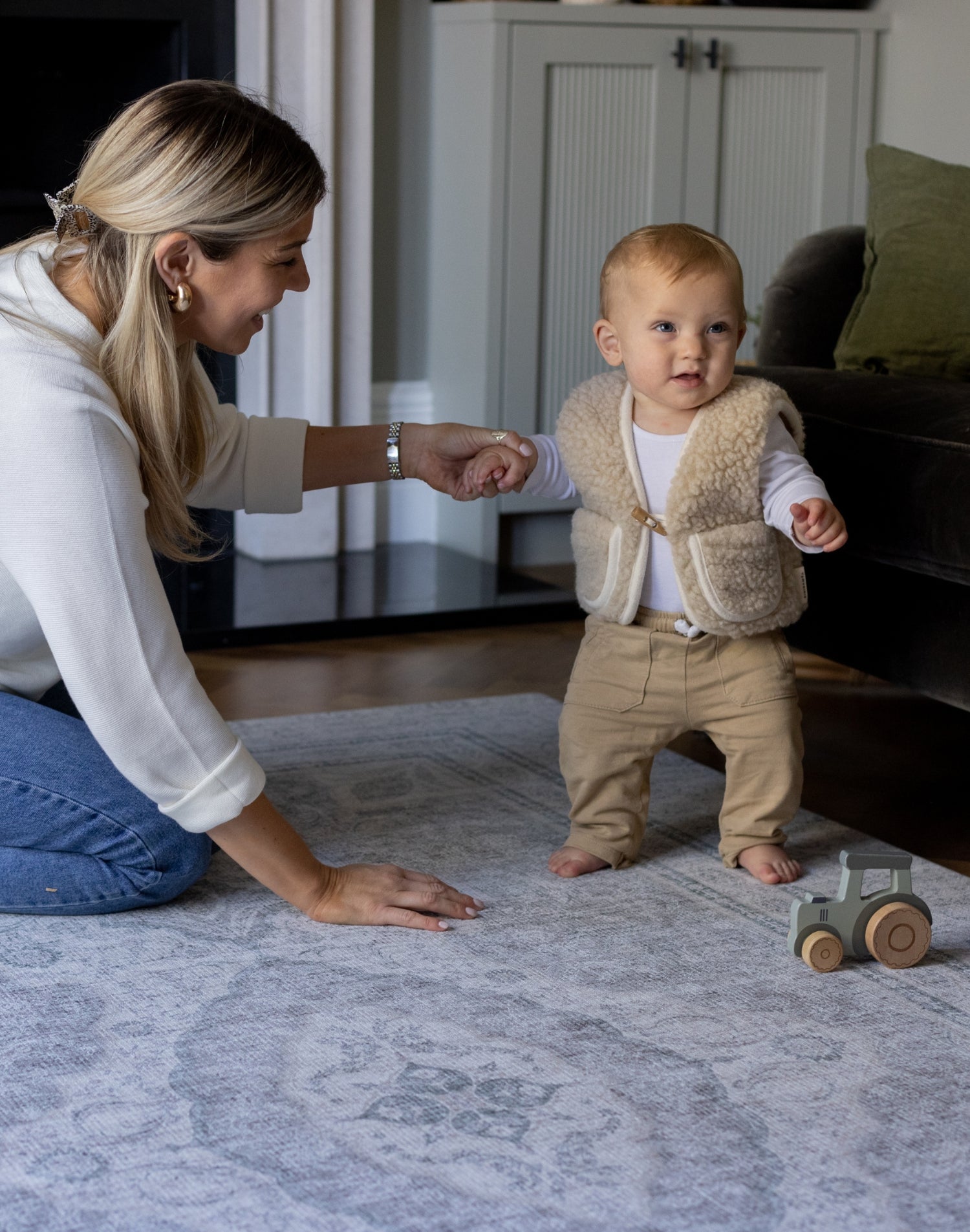 A mother holds the hand of a baby who is learning to walk. The baby is walking on a big soft foam playmat that has a beautiful grey design