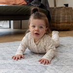 Baby crawls doing tummy time on a large waterproof play mat. The background is a family home interior space