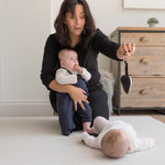 Lady holding one baby and playing with a second baby laying on a thick foam play mat that supports children and adults on the floor